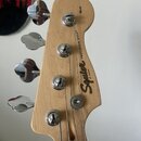 Hlava basové kytary z přírodního světlého dřeva s chromovanými ladičkami. Na dřevě je černý nápis Squier BY FENDER a PRECISION BASS®. V horní části h…