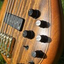 Detail těla basové kytary s přírodním dřevem a viditelnou sletovkou. Nástroj má dva černé snímače, na kterých je bílé logo ve tvaru stylizovaného pís…