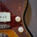 Detail elektrické kytary s žlutohnatým průsvitným povrchem a červeně hnědým želvovinovým pickguardem. Na knoflících jsou nápisy VOLUME a TONE s číslo…