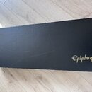 Černý obdélníkový kufr na hudební nástroj s texturovaným povrchem. V pravé dolní části je zlatý nápis Epiphone. Povrch vykazuje drobné škrábance a zn…