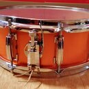 Pomalu vyfotocený oranžový snare drum s chromovaným obručováním a mechanismem. Na koruse nejsou viditelné žádné značky ani nápisy. Povrch nástroje vy…