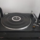 Černý gramofon s černou gumičkou na talíři a stříbrným vnějším okrajem. Vpravo je umístěno raménko s hloubkou a jehla. Na spodní části přístroje jsou…