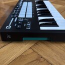 Černý MIDI kontrolér s klávesnicí a bílými pady. Na levé straně je bílé logo novation a nápisy TRACK, OCTAVE, Hold, MIDI Channel. V zadní části je vi…