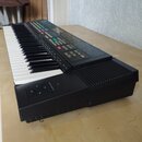 Černý elektronický klávesový nástroj s bílými a černými klávesami. Horní panel obsahuje displej, řadu barevných tlačítek a ovládacích prvků. V levé č…