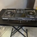 Černý DJ 컨트롤러 Numark MIXSTREAM PRO umístěný na černém stole. Na přední straně je vidět bílý nápis Numark MIXSTREAM PRO a menší text pod ním, který je…