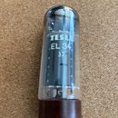 Fotografie znázorňuje elektronovou lampu s prohladněným skleněným krytem a hnědým plastovým základním soketem. Na skle je bílý nápis TESLA, EL 34 a n…