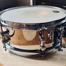 Chromovaný snare drum s bílým blátcem. Na blátci je viditelné logo značky REMO. Nástroj vykazuje známky běžného používání v podobě drobných znečištěn…