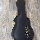 Černý tvrdý kufr na kytaru s zlatými zapojovacími prvky. Na povrchu je zlatý nápis Epiphone a logo Gibson Custom Shop v kosočtverci. Povrch kufru vyp…