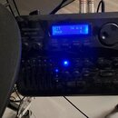 Černý modul elektronických bicích s modrým podsvícením displeje. Na levé straně je bílý nápis Millenium a pod ním model MPS-750X. Displej zobrazuje t…