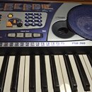 Stříbrný elektronický klavír s černými a bílými klávesami. Na ovládacím panelu je nápis PSR-260, logo YAMAHA a označení BASS BOOST SYSTEM. Jsou zde v…