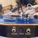 Detailní záběr na spodní hranu modré kytary s viditelným žlutým lemem. Na černé ovládací desce jsou tři výstupní konektory s nápisy Mix, Acoustic a E…
