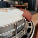 Detailní pohled na horní hranu bílého bubnu s chromovaným obručem. Na okraji je připevněna hnědá, zaoblená leather-pad podložka z kůže nebo syntetick…