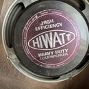 Fotografie zachycuje zadní část reproduktoru s kovovým rámem. Na středovém magnetu je purpurový štít s bílým nápisem HIWATT, nad kterým je text HIGH…