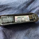 Černý oválný plastový modul s vnitřním zapojením. Na bílém štítku je uveden nápis EMG-SLV, MADE IN USA G 1710.01, (21)4870185 a datum 07/09/2026 spol…