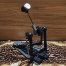 Černý basový pedál s kovovou konstrukcí a řetězovým pohonem. Na šlapadle je vidět vyfrézovaný vzorek a na horní části hracího mechanismu je bílá pols…