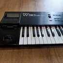 Černý digitální klávesový nástroj s bílými a černými klávesami. Na horním panelu je bílý nápis Roland W-30 MUSIC WORKSTATION. Vlevo se nachází disket…