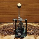 Černý bubnový pedál s kovovými mechanismy a světlým, kulatým nášlapem. Na základní desce je vidět nápis "TAMA". Povrch nástroje vykazuje mírné známky…