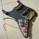 Černý plastový pickguard s dvěma snímači a zlatým kovovým mostkem. V dolní části je vidět rozvedená elektronika s potenciometry a přepínačem na bílém…
