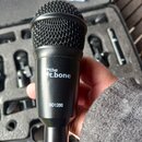 Černý dynamický mikrofon s kovovou mřížkou. Na těle je bílý nápis „the t.bone“ a níže označení modelu „BD1200“. Mikrofon je v ruky osoby, v pozadí je…