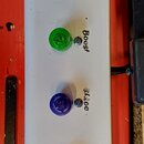 Bílý obdélníkový pedalboard s čtyřmi barevnými tlačítky: modrým, zeleným, fialovým a oranžovým. Vedle tlačítek jsou nápisy v kurzívě: „Channel“, „Boo…