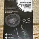 Fotografie znázorňuje krabici s produktem SOUNDHOLE MICROPHONE & WIRELESS SYSTEM značky ORTEGA GUITARS • EST 1994. Na obalu je zobrazen černý kulatý…