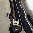 Černá elektrická basová kytara v otevřeném tvrdém kufru s černým vnitřkem. Na hlavě nástroje je viditelné logo Fender. Kytara má bílý pickguard, čtyř…