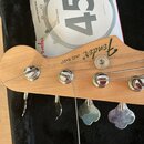 Detail hlavy basové kytary z přírodního dřeva s nápisy Fender® a Jazz BASS. Na hlavě jsou čtyři chromované ladičky s kovovými knoflíky. V pozadí je v…