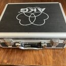Černý přepravní kufr s hliníkovými hranami a kovovými zámky. Na víku je bílé logo AKG s doprovodným symbolem. Na přední straně u rukojeti je nápis AK…