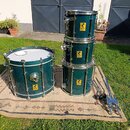 Sada bubnů v tmavě zelené barvě s kovovým hardwarem. Na každém z bubnů je žlutý čtvercový štítek s nápisem „FIRE SHELL“ a symbolem radioactivity. Sad…