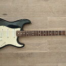 Černá elektrická kytara s bílým pickguardem a hnědým hmatníkem. Na hlavě je viditelné logo Fender a nápis STRATOCASTER. Kytara má tři snímače, pět úh…