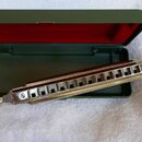 Stříbrná harmonika s kovovým povrchem a řadou otvorů, umístěná před otevřeným zeleným kovovým pouzdrem s červeným vnitřkem víka. Na nástroji nejsou v…