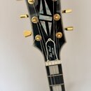 Fotografie zachycuje hlavu a horní část hmatníku černé elektrické kytary. Na hlavě je bílý nápis Epiphone a níže štítek s textem Les Paul CUSTOM. Nás…