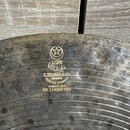 Detailní záběr na povrch kovového činele s viditelným opotřebením a oxidací. Na povrchu je zlatý nápis s logem hvězdy v kruhu, arabským písmem a text…
