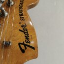 Detail hlavy kytary z dřeva s lakovaným povrchem. Na hlavě je černý nápis Fender® STRATOCASTER a malé vertikální sériové číslo 3143028. Jsou vidět ko…