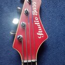 Detail hlavy červené basové kytary s bílým lemováním. Na povrchu je bílý nápis „Studio Bass“. Jsou vidět čtyři kovové ladičky a struny, z nichž jedna…
