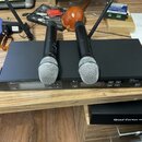 Černý rackový přijímač s dvěma bezdrátovými mikrofony s kovovou mřížkou. Na přední straně přijímače je nápis Micro Lcbone free solo Tex (200 MHz). Po…