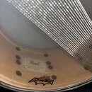 Detail vnitřku bubnu s napnutou blánou značky REMO, na které je nápis AMBASSADOR SNARE HEAD. Uprostřed blány je nalepená štítková nálepka s logem Pea…