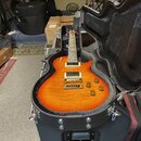 Elektrická kytara s trupem z plamenového javoru v oranžovém přechodovém odstínu, umístěná v černém tvrdém kufru. Nástroj má dva humbuckery a čtyři ov…