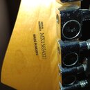 Detail zadní strany hlavy kytary z jasného dřeva. Je zde černý nápis SERIAL NUMBER MX15654327 a pod ním MADE IN MEXICO. Vpravo jsou vidět tři kovové…