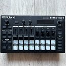 Černá groovebox s nápisy Roland a GROOVEBOX MC-101. Zařízení obsahuje displej, několik otočných ovladačů a bílá tlačítka s různými označením, včetně…