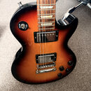 Elektrická kytara s tmelelovým povrchem v přechodové barvě od černé po žlutohnědou. Nástroj má dva humbuckery, čtyři ovládací knofíky a přepínač s ná…