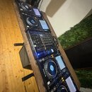 Sada DJského vybavení na dřevěném stole, zahrnující čtyři přehrávače a jeden mixážní pult. Na zařízeních je vidět nápis Pioneer DJ. Všechny komponent…
