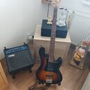 Elektrická basová kytara s tmavě hnědým sunburst povrchem a hmatníkem z tmavého dřeva. Na hlavě nástroje je bílý nápis Cort. Vedle kytary stojí malý…