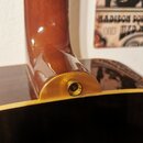Detailní pohled na spojení krku a korpusu akustické kytary v hnědé barvě s lakovaným povrchem. V oblasti paty krku je umístěn kovový výstup pro jack…