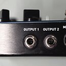Detail zadní strany černého elektronického zařízení s kovovým, broušeným panelem. Jsou zde vidět tři konektory s nápisy OUTPUT 1, OUTPUT 2 a MIDI THR…