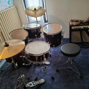 Černá bicí souprava s kovovými obručemi, která obsahuje tři bubny a dva činely. Na blanách bubnů je viditelný nápis Multibeat. Souprava je doplněna c…