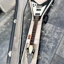 Černá elektrická kytara s bílým pickguardem v tvaru Flying V, umístěná v černém tvrdém kufru s bílým vnitřkem. Nástroj má hnědý hmatník a stříbrné po…