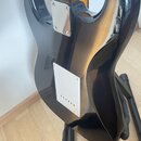 Zadní strana černé elektrické kytary s lesklým povrchem, umístěná na černém stojanu. Na těle jsou vidět dva kovové kryty: horní čtvercový s čtyřmi šr…