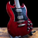Elektrická kytara s červeným lakem a černým pickguardem, umístěná na černém stojanu. Nástroj má dva humbuckery a čtyři ovládací knoflíky. Povrch laku…