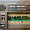 Šedý elektronický klavír značky Roland s otevřeným vnitřkem, kde je viditelná zelená deska plošných spojů s nápisem 052 H 482-1. Na ovládacím panelu…